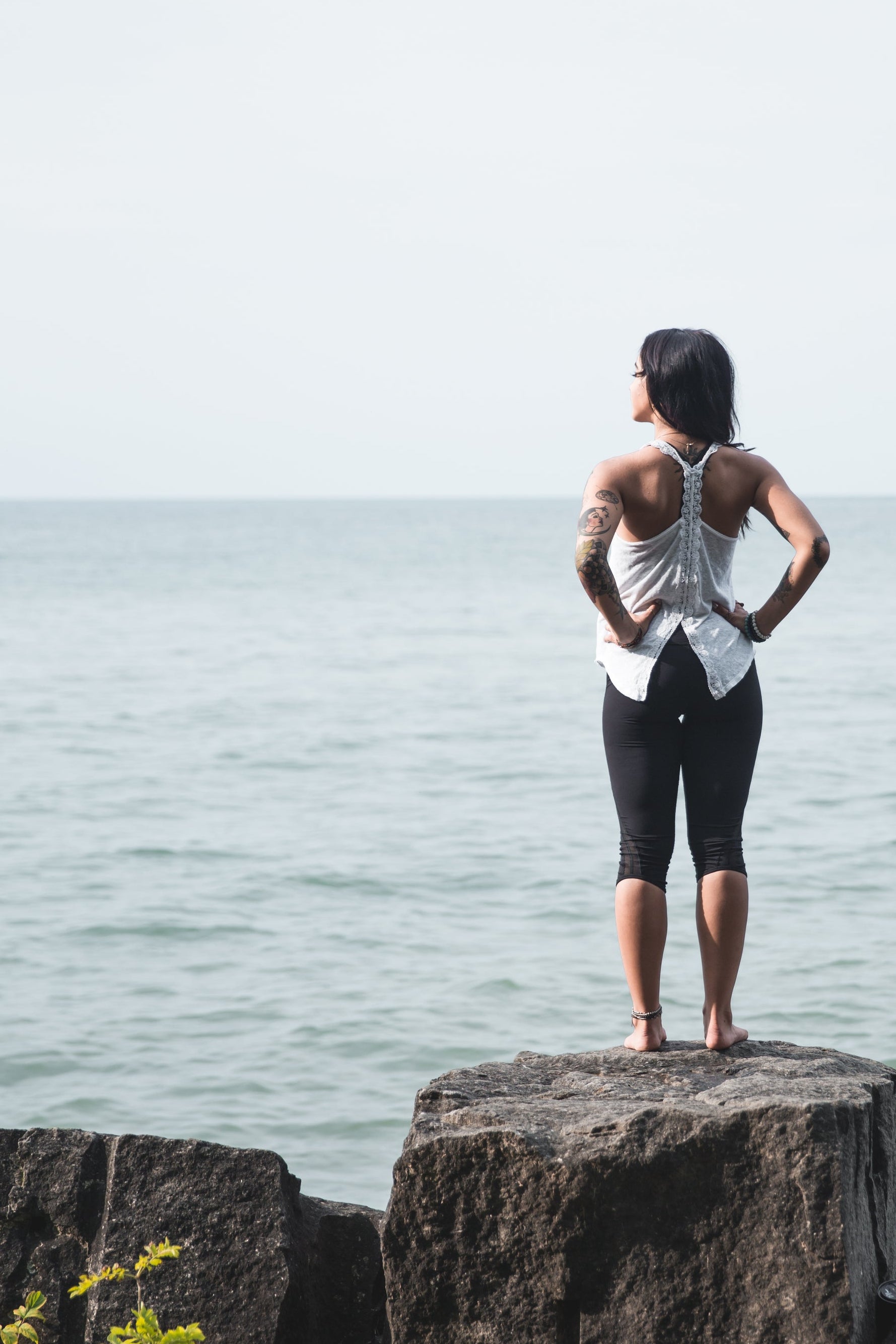 Person standing on a rock by the ocean, wearing a white sleeveless top and black shorts.